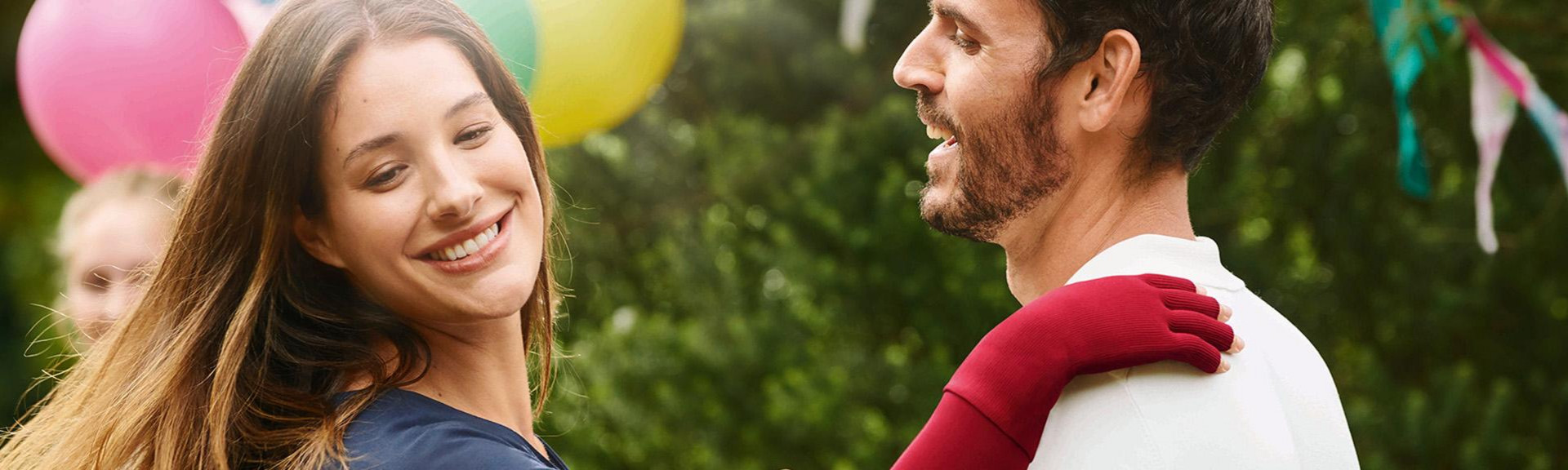 Close up of couple dancing in the garden with balloons in the background, both smiling, woman wearing compression on hand which is on partner's shoulder