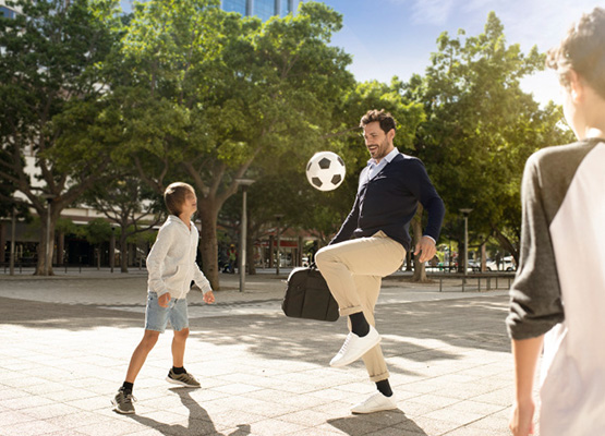 Dad playing football wearing office work wear and holding briefcase kicking a football in park with his sons