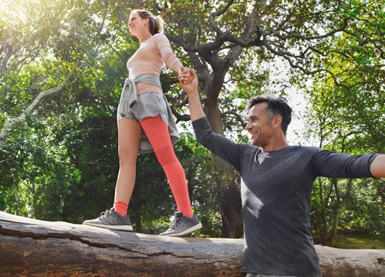 Women wearing compression on her leg walking across a log in woodland holding male's hand