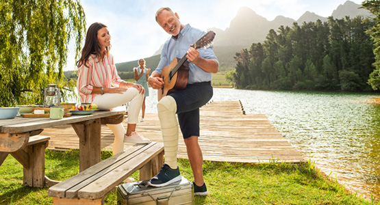 Man playing his guitar stood up with his leg on a wooden bench beside a lake with his wife next to him